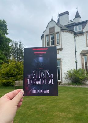 A picture of a white hand holding a postcard that reads "The Ghosts of Thorwald Place" in front of a
