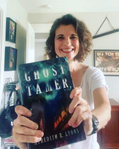 A white woman with short brown hair smiling at the camera while holding up the hardcover book "Ghost Tamer" by Meredith R. Lyons. The background is a comfy living room