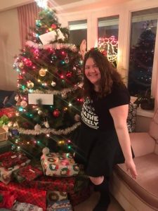 Maddie dressed in a black Christmas T-Shirt and a black shirt posing beside the Christmas tree with a large pile of presents