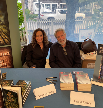 Picture of Marcy and her husband, Lew, at a table with stacks of both of their books and pens