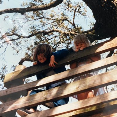 A picture of two young girls smiling down at the camera over a wooden fence-like structure, with a tree in and bright blue sky in the background