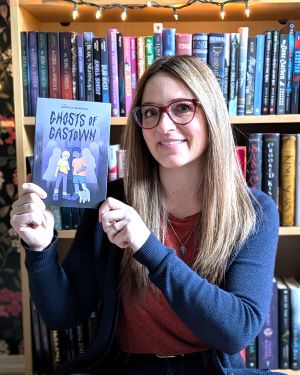 A white woman with long, light brown hair smiling at the camera holding up a copy of the book "Ghosts of Gastown", and sitting in front of bookshelves with lights